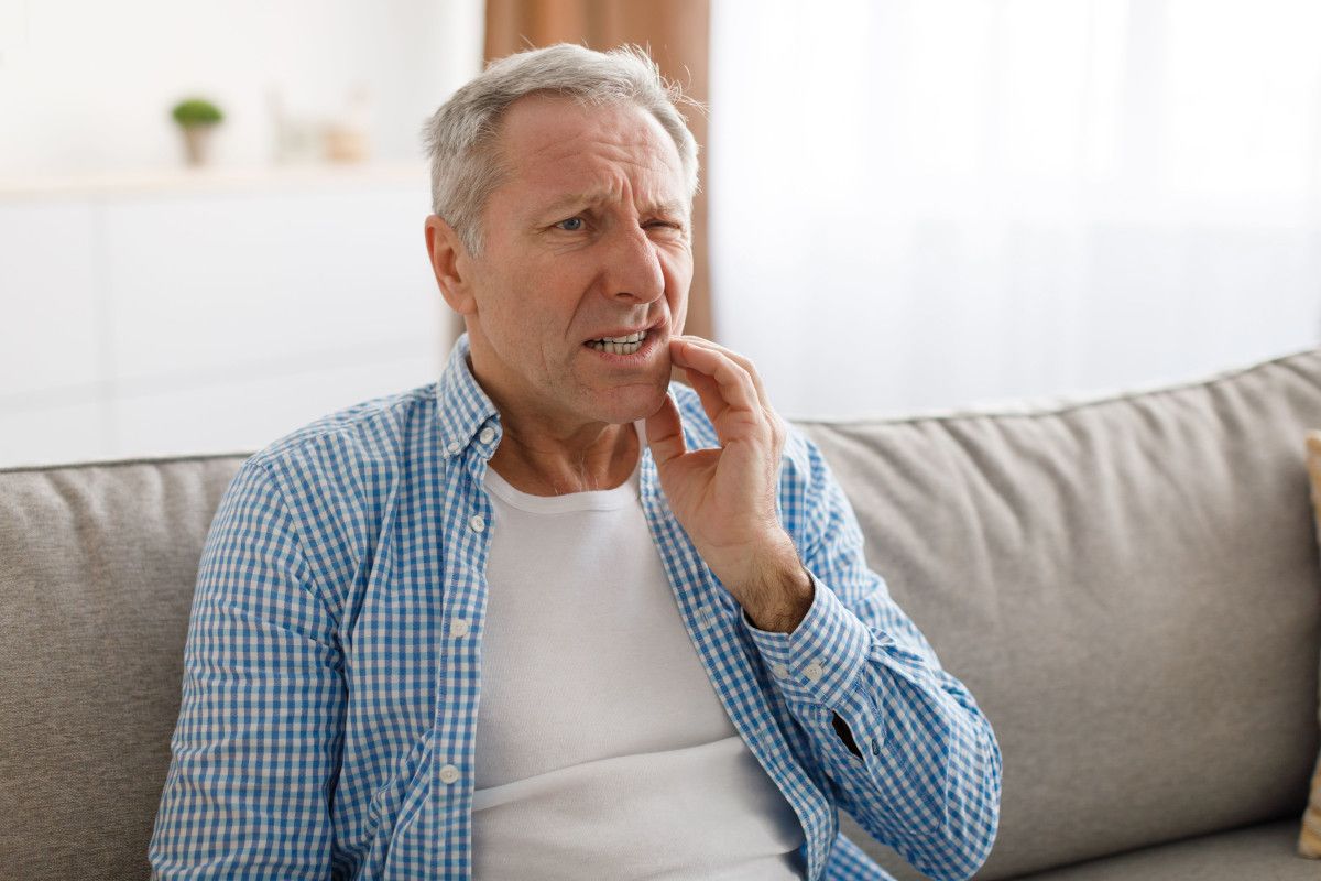 How to Handle a Dental Emergency Mature man sitting on couch, hand to face, appearing thoughtful or experiencing tooth pain.